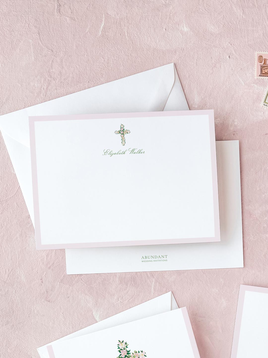 White flat notecard with a delicate watercolor floral cross design and light pink border on a textured pink background