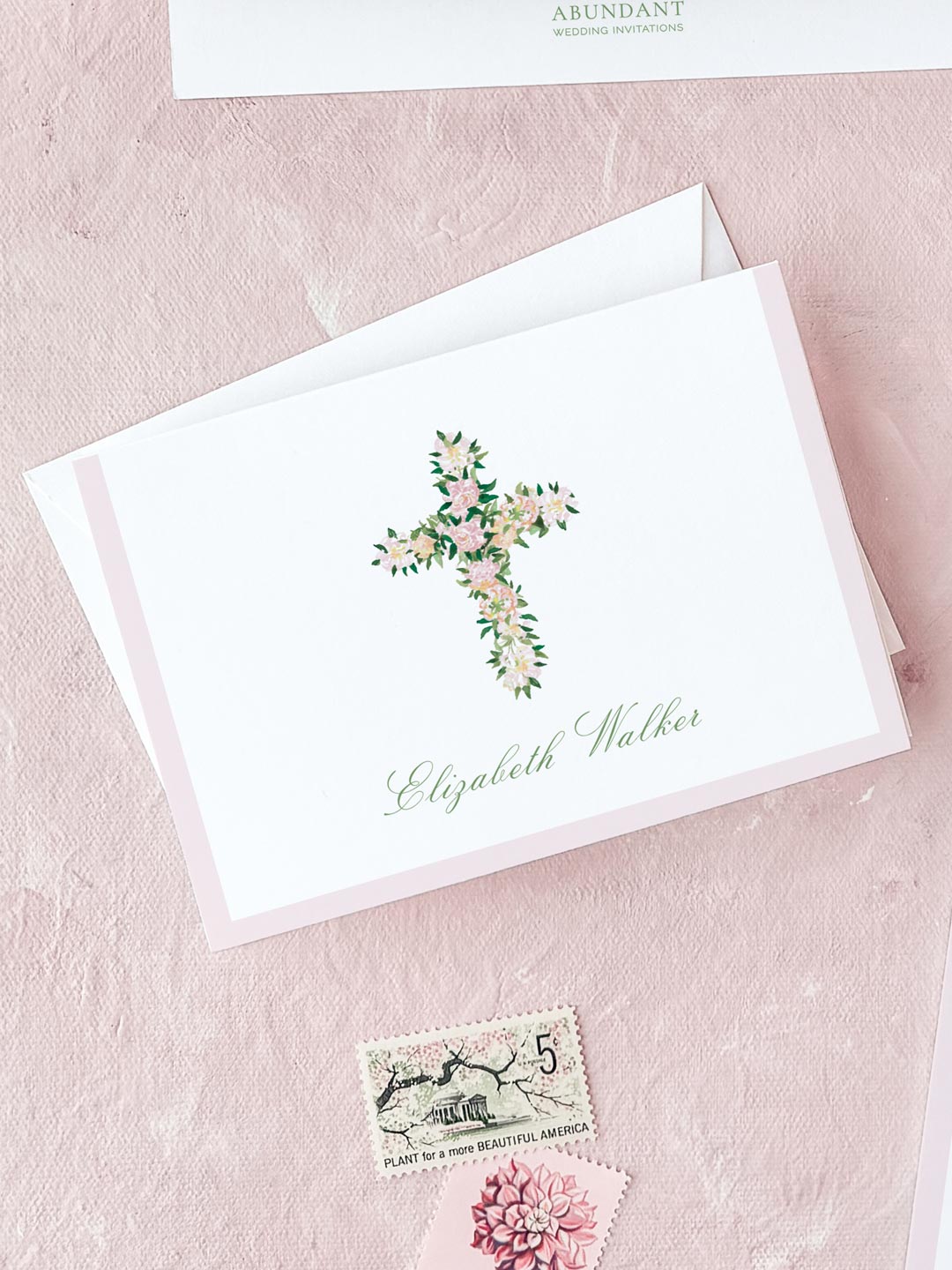 Folded notecard with a watercolor floral cross and elegant script on soft pink textured background.