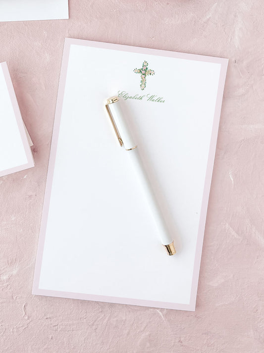 White notepad with floral cross design and elegant pen on a soft pink textured surface