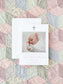Birth announcement card featuring a soft watercolor floral cross and a photo of a sleeping newborn on a pastel patchwork quilt background.