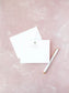 Elegant flat notecard with a delicate watercolor cross and soft gray text, laying next to a matching envelope and white pen on a textured pink background.