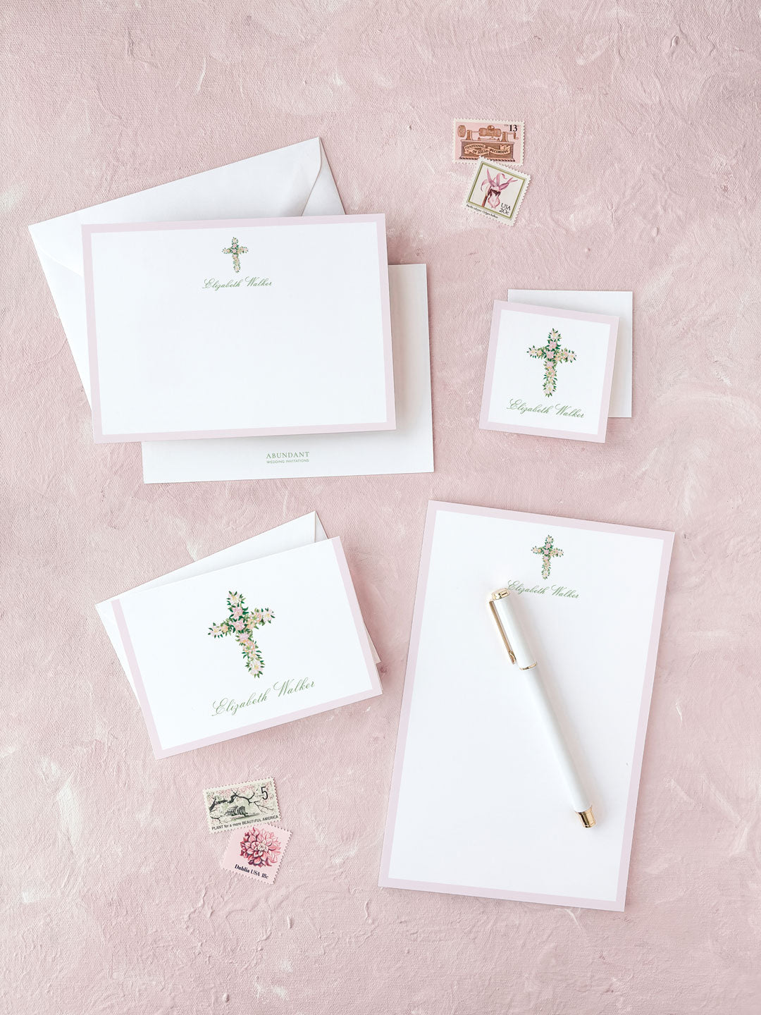 Stationery set with floral cross design, including notepad, cards, and envelopes on a pink textured surface.
