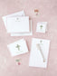 Stationery set with floral cross design, including notepad, cards, and envelopes on a pink textured surface.