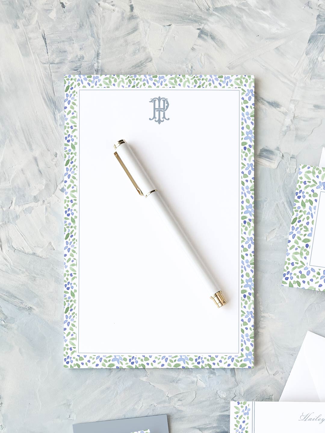 Elegant notepad with blue and green watercolor floral border and a white pen on textured surface